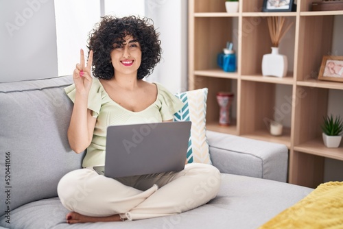 Young brunette woman with curly hair using laptop sitting on the sofa at home showing and pointing up with fingers number two while smiling confident and happy.