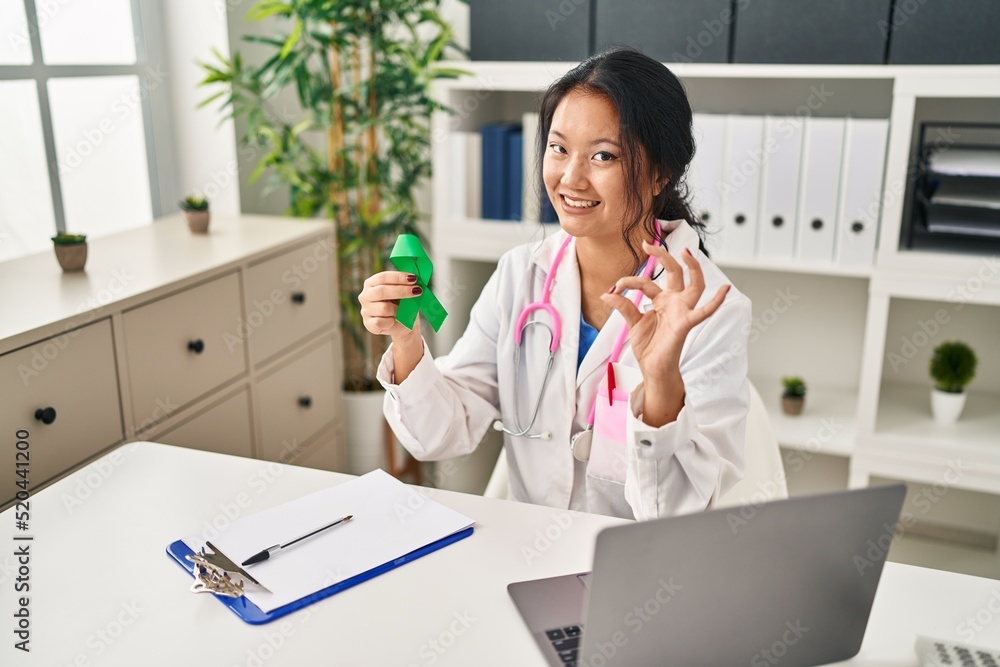 Young asian doctor woman holding support green ribbon doing ok sign with fingers, smiling friendly gesturing excellent symbol