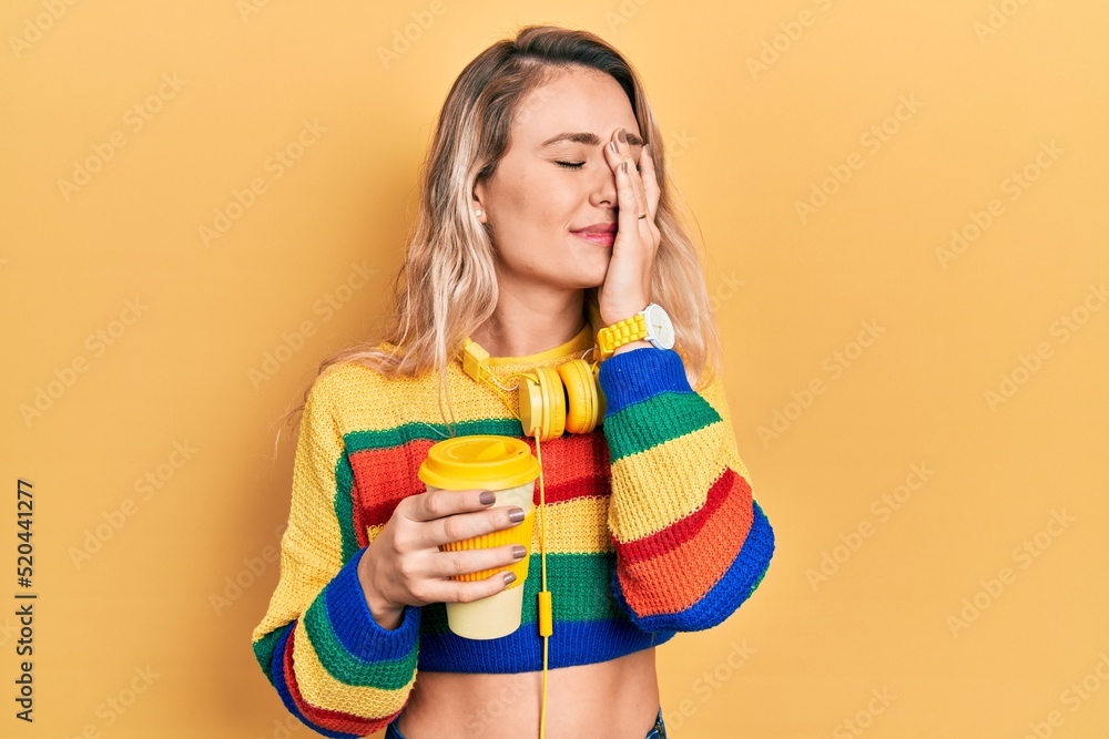 Beautiful young blonde woman drinking cup of coffee wearing headphones yawning tired covering half face, eye and mouth with hand. face hurts in pain.