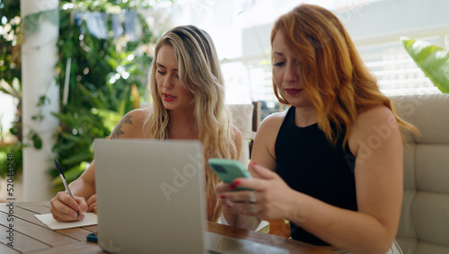 Two women using smartphone writing on notebook sitting on table at home terrace