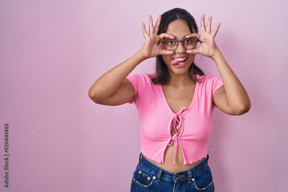Hispanic young woman standing over pink background wearing glasses doing ok gesture like binoculars sticking tongue out, eyes looking through fingers. crazy expression. Hispanic young woman standing over pink background wearing glasses doing ok gesture like binoculars sticking tongue out, eyes looking through fingers. crazy expression.