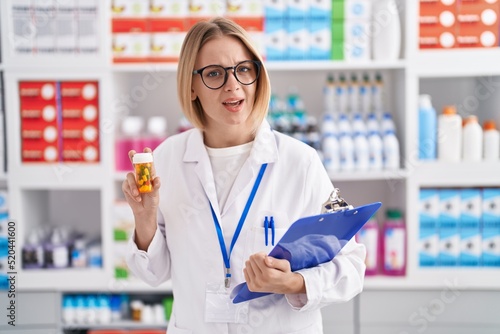 Obraz na plátně Young caucasian woman working at pharmacy drugstore holding pills clueless and confused expression
