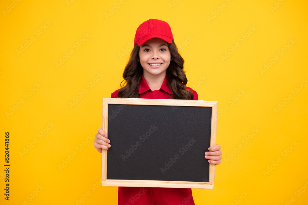 Child girl builder in red building uniform and cap. Teenage girl worker hold blackboard isolated on yellow background. Kids renovation concept. Copy space on empty board, mock up for menu. Child girl builder in red building uniform and cap. Teenage girl worker hold blackboard isolated on yellow background. Kids renovation concept. Copy space on empty board, mock up for menu.