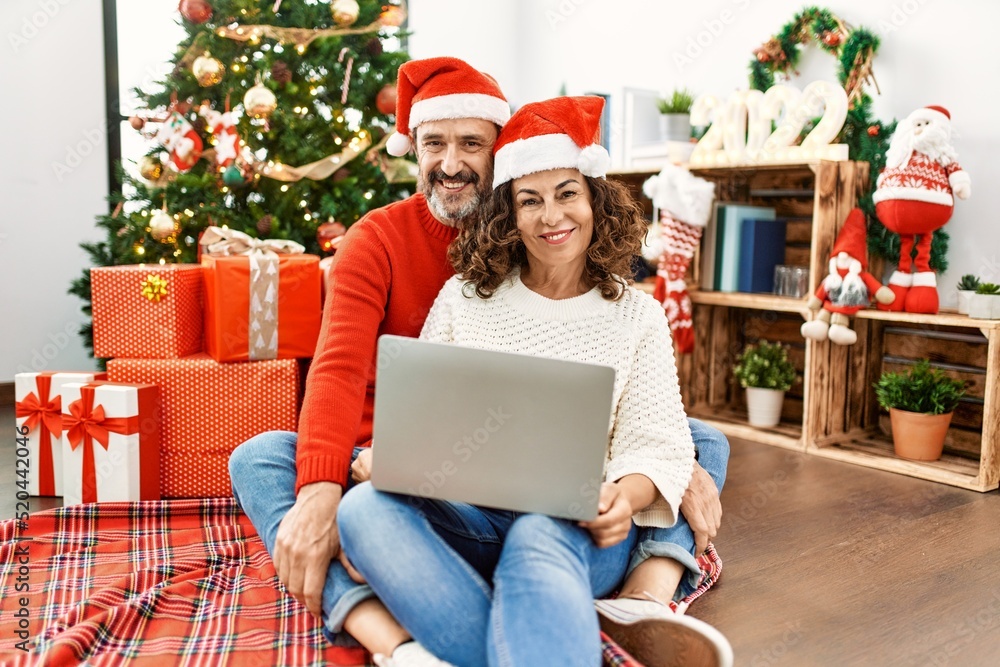 Hispanic middle age woman and mature man using laptop sitting by christmas tree looking positive and happy standing and smiling with a confident smile showing teeth Hispanic middle age woman and mature man using laptop sitting by christmas tree looking positive and happy standing and smiling with a confident smile showing teeth