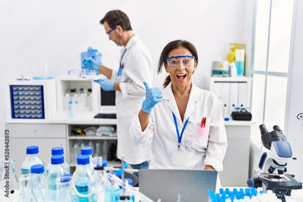 Middle age woman working at scientist laboratory pointing thumb up to the side smiling happy with open mouth Middle age woman working at scientist laboratory pointing thumb up to the side smiling happy with open mouth