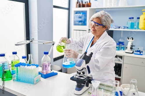 Middle age woman wearing scientist uniform pouring liquid on test tube at laboratory