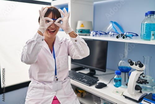 Young brunette woman working at scientist laboratory doing ok gesture like binoculars sticking tongue out  eyes looking through fingers. crazy expression.