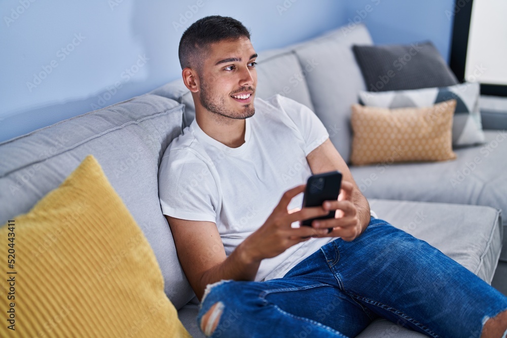Young hispanic man using smartphone sitting on sofa at home Young hispanic man using smartphone sitting on sofa at home