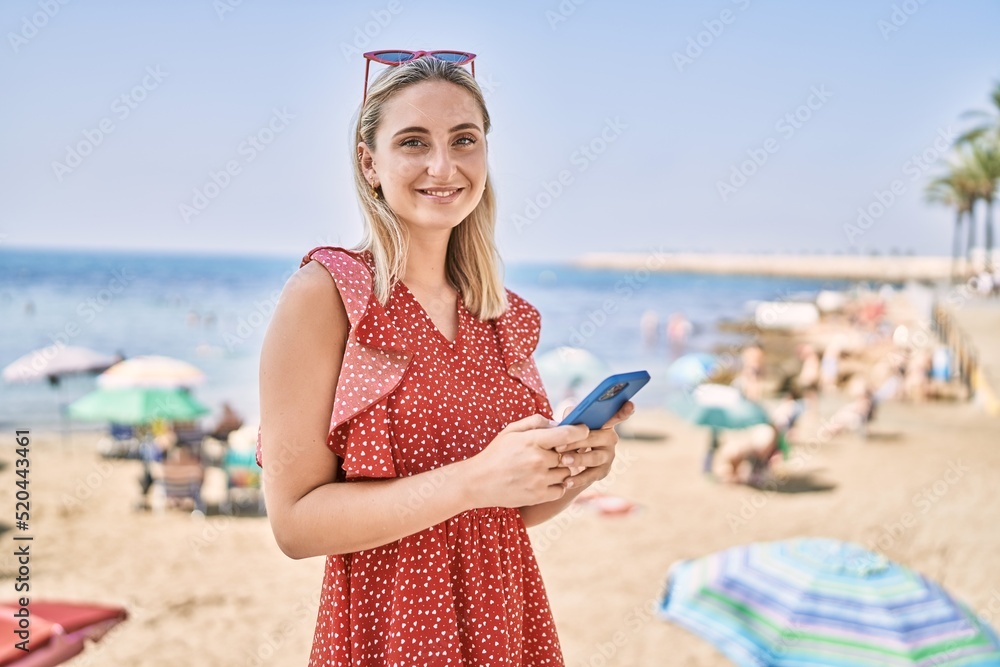 Young blonde girl smiling happy using smartphone at the beach