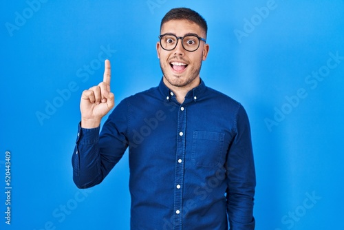 Young hispanic man wearing glasses over blue background pointing finger up with successful idea. exited and happy. number one.