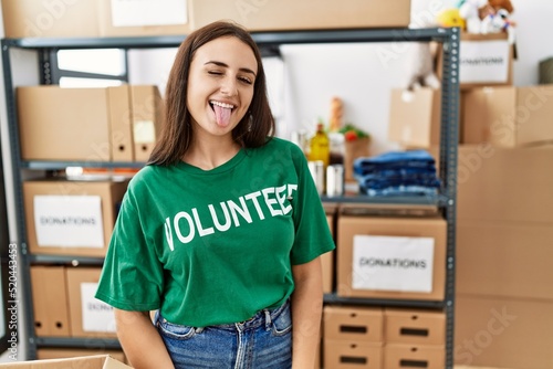 Young brunette woman wearing volunteer t shirt at donations stand sticking tongue out happy with funny expression. emotion concept.