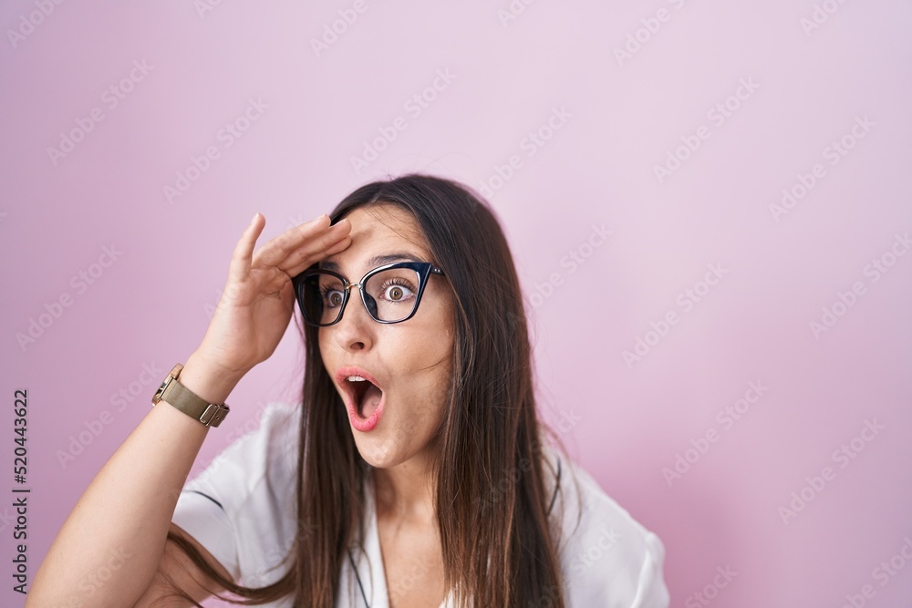 Young brunette woman wearing glasses standing over pink background very happy and smiling looking far away with hand over head. searching concept. Young brunette woman wearing glasses standing over pink background very happy and smiling looking far away with hand over head. searching concept.