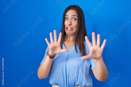 Картината върху платно Young brunette woman standing over blue background afraid and terrified with fear expression stop gesture with hands, shouting in shock