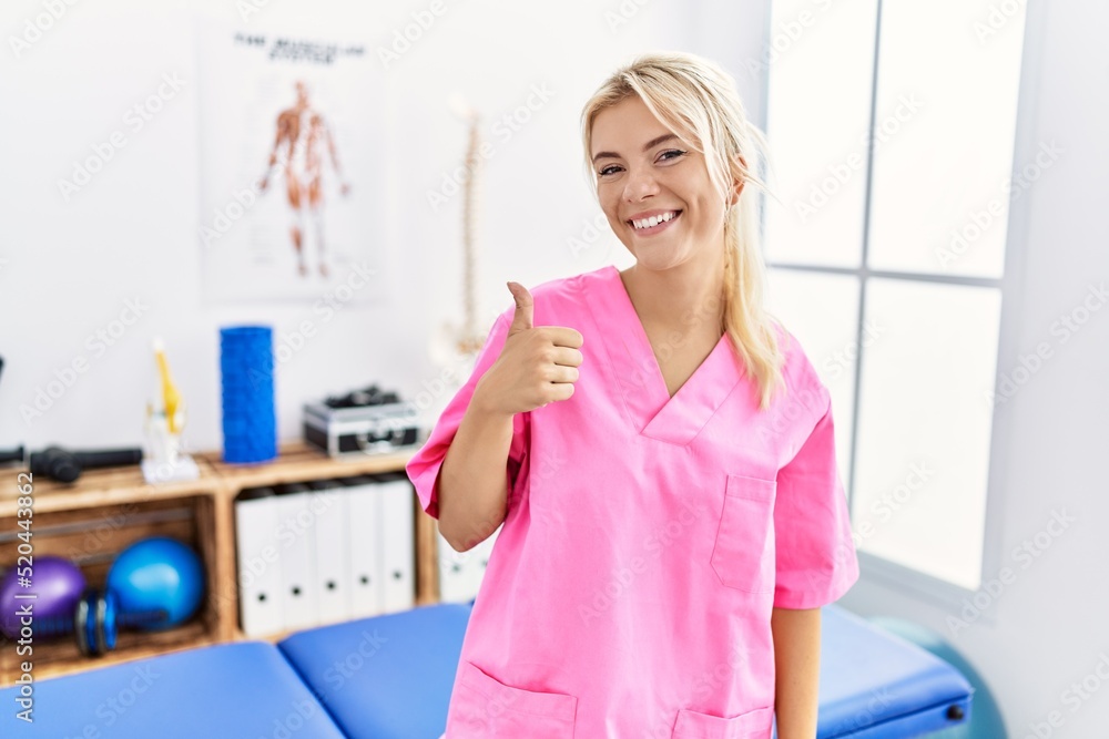 Young caucasian woman working at pain recovery clinic doing happy thumbs up gesture with hand. approving expression looking at the camera showing success.