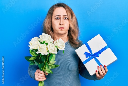 Middle age caucasian woman holding anniversary present and bouquet of flowers puffing cheeks with funny face. mouth inflated with air, catching air.