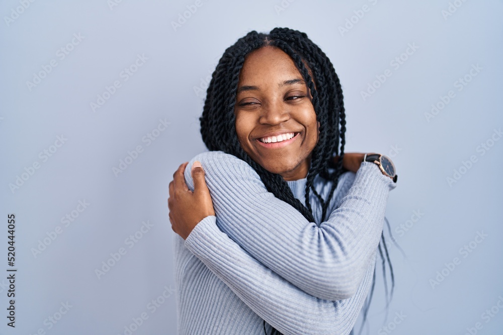 African american woman standing over blue background hugging oneself happy and positive, smiling confident. self love and self care African american woman standing over blue background hugging oneself happy and positive, smiling confident. self love and self care
