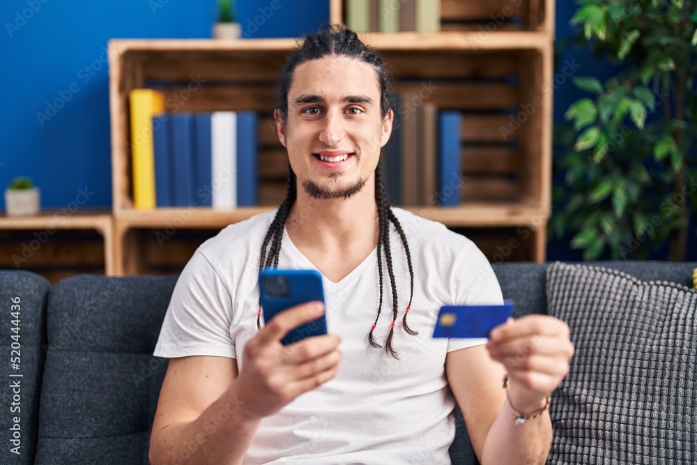 Young man using smartphone and credit card sitting on sofa at home