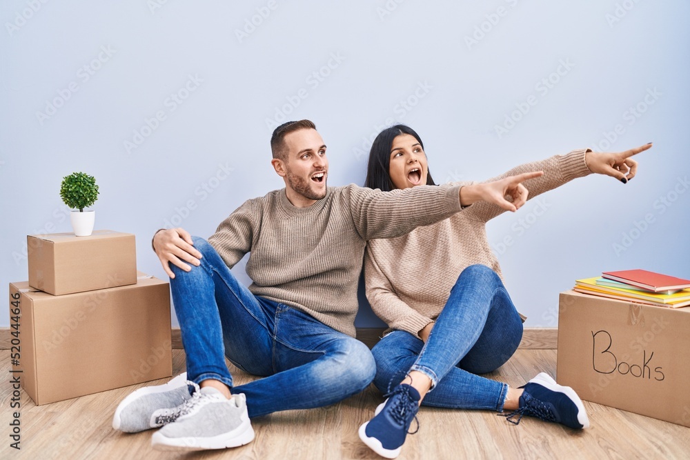Young couple moving to a new home pointing with finger surprised ahead, open mouth amazed expression, something on the front Young couple moving to a new home pointing with finger surprised ahead, open mouth amazed expression, something on the front