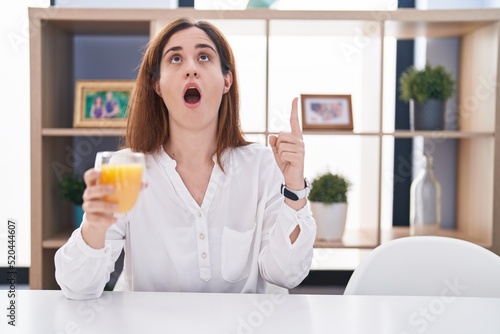Brunette woman drinking glass of orange juice amazed and surprised looking up and pointing with fingers and raised arms.