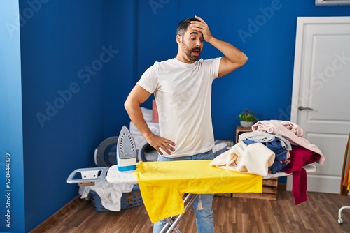 Young hispanic man with surprise expression looking clothes at laundry room