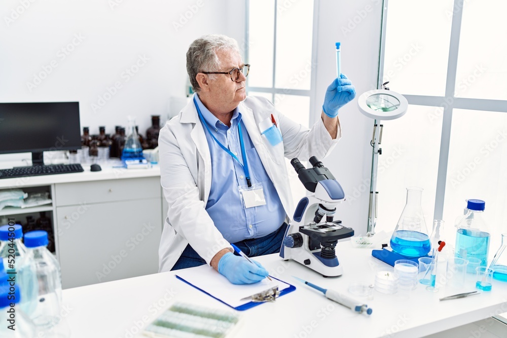 Middle age grey-haired man wearing scientist uniform holding test tube and writing on clipboard at laboratory Middle age grey-haired man wearing scientist uniform holding test tube and writing on clipboard at laboratory