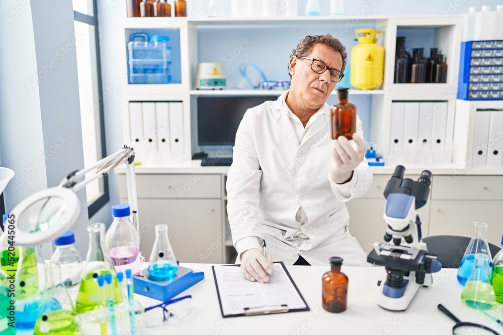 Middle age man wearing scientist uniform holding bottle writing on clipboard at laboratory Middle age man wearing scientist uniform holding bottle writing on clipboard at laboratory