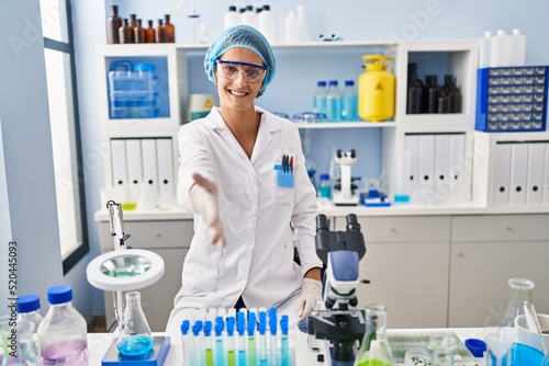 Brunette woman working at scientist laboratory smiling friendly offering handshake as greeting and welcoming. successful business.