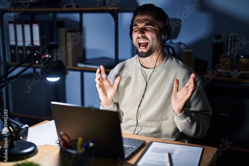Young handsome man working using computer laptop at night crazy and mad shouting and yelling with aggressive expression and arms raised. frustration concept.