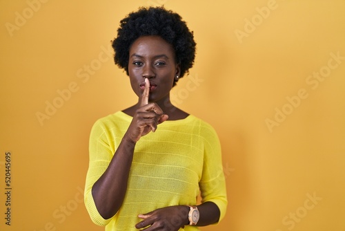 African young woman standing over yellow studio asking to be quiet with finger on lips. silence and secret concept. © Krakenimages.com