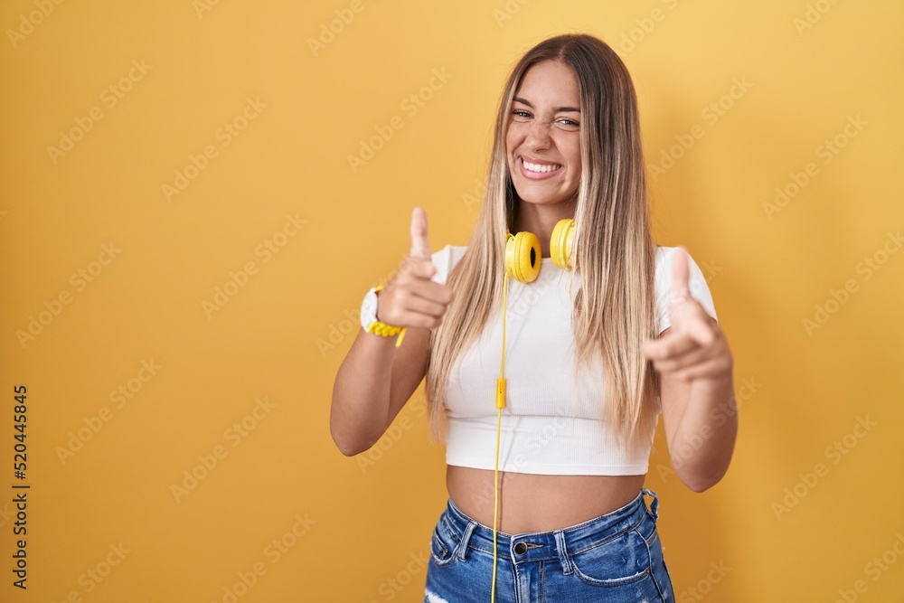 Young blonde woman standing over yellow background wearing headphones pointing fingers to camera with happy and funny face. good energy and vibes. Young blonde woman standing over yellow background wearing headphones pointing fingers to camera with happy and funny face. good energy and vibes.