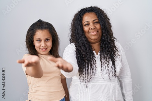Mother and young daughter standing over white background smiling cheerful offering palm hand giving assistance and acceptance.