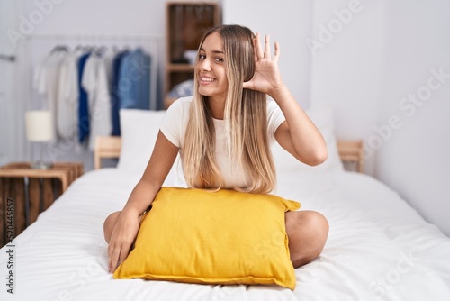 Young blonde woman sitting on the bed with pillow at home smiling with hand over ear listening an hearing to rumor or gossip. deafness concept.
