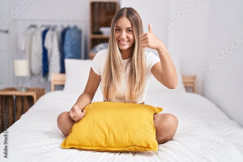 Young blonde woman sitting on the bed with pillow at home doing happy thumbs up gesture with hand. approving expression looking at the camera showing success.