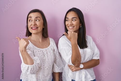 Hispanic mother and daughter together smiling with happy face looking and pointing to the side with thumb up.