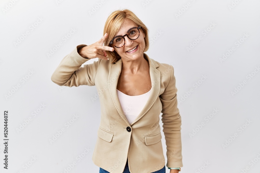 Middle age blonde business woman standing over isolated background doing peace symbol with fingers over face, smiling cheerful showing victory Middle age blonde business woman standing over isolated background doing peace symbol with fingers over face, smiling cheerful showing victory