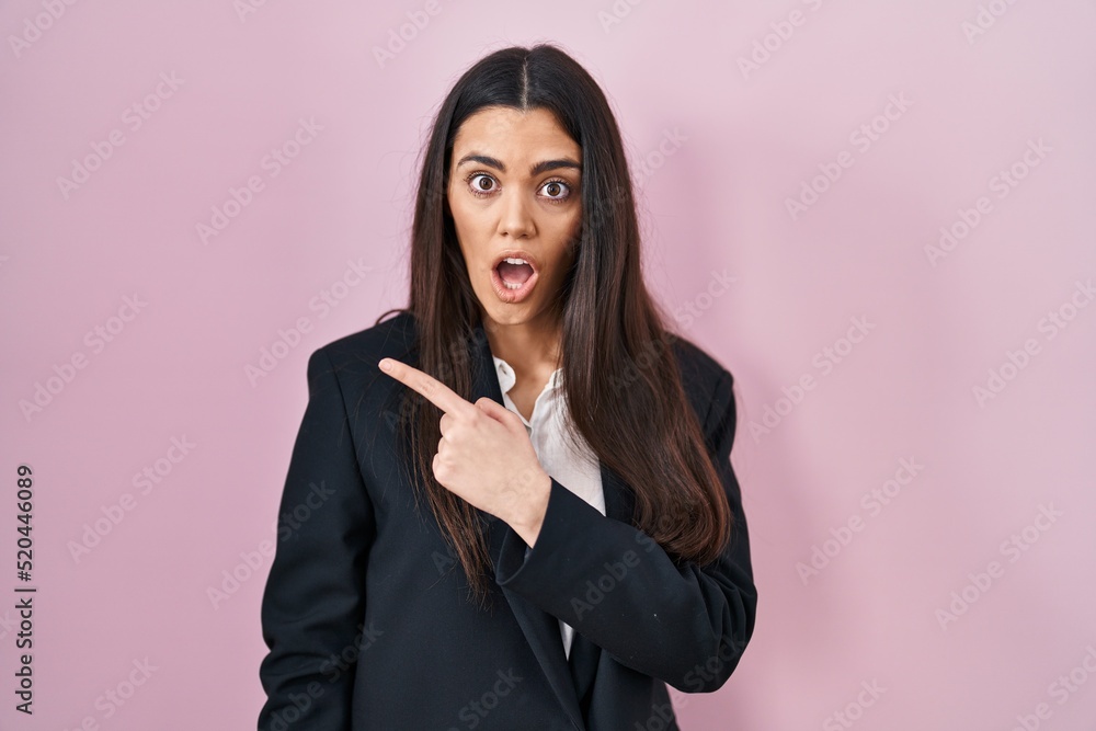 Young brunette woman wearing business style over pink background surprised pointing with finger to the side, open mouth amazed expression. Young brunette woman wearing business style over pink background surprised pointing with finger to the side, open mouth amazed expression.