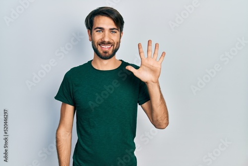Young hispanic man wearing casual white tshirt showing and pointing up with fingers number five while smiling confident and happy.