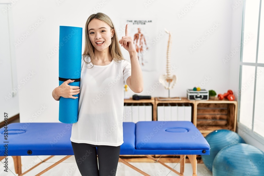 Asian young woman holding yoga mat at pain recovery clinic surprised with an idea or question pointing finger with happy face, number one