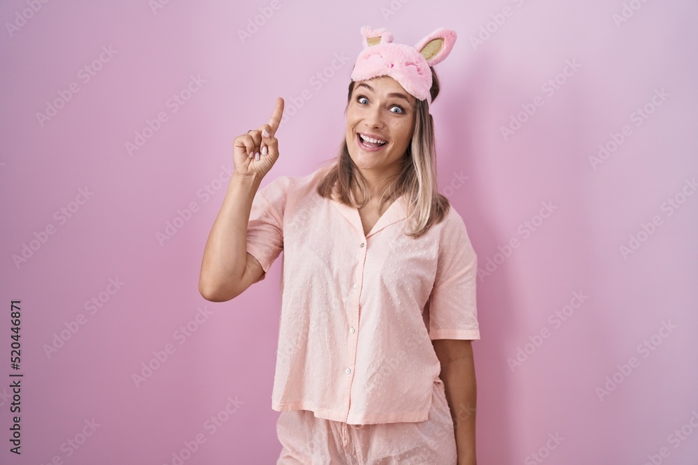 Blonde caucasian woman wearing sleep mask and pajama pointing finger up with successful idea. exited and happy. number one. Blonde caucasian woman wearing sleep mask and pajama pointing finger up with successful idea. exited and happy. number one.