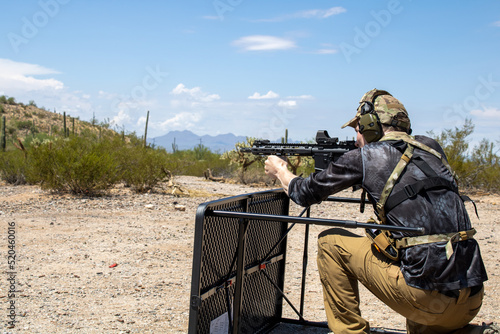 Male holding ar15 pistol with vertical foregrip and holosight and wearing ear protection while taking cover behind table and shooting targets