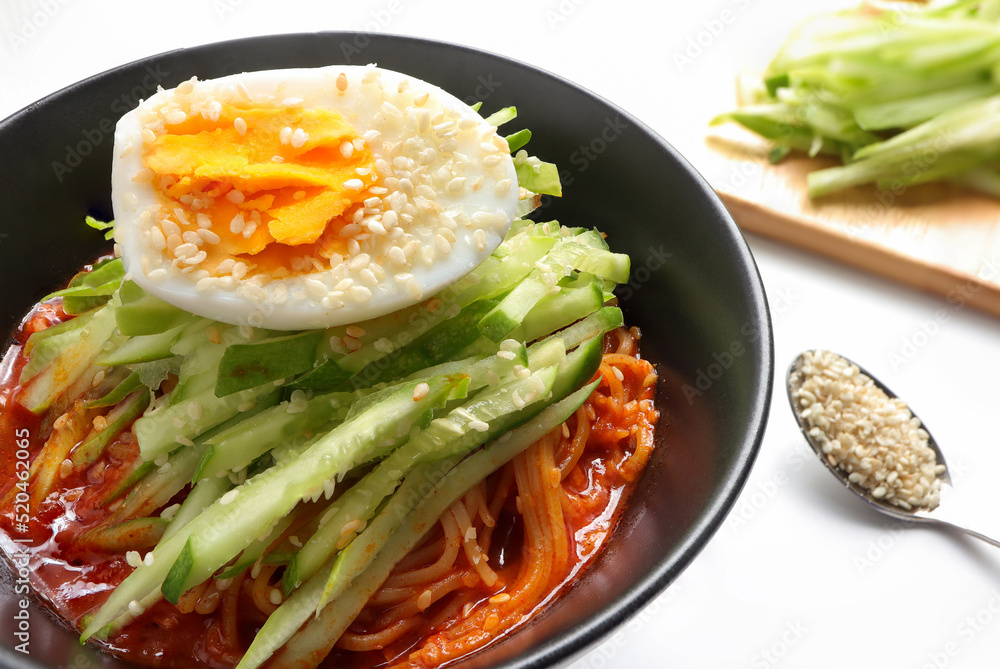 Korean cold noodles with spicy sauce, topped with cucumber, boiled egg and crushed sesame seeds in a black bowl. Bibim Guksu or Korean cold noodle salad (Spicy mixed noodles) Usually eat in Summer. Korean cold noodles with spicy sauce, topped with cucumber, boiled egg and crushed sesame seeds in a black bowl. Bibim Guksu or Korean cold noodle salad (Spicy mixed noodles) Usually eat in Summer.
