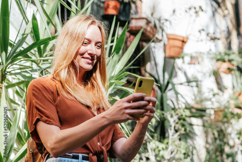 cheerful blonde female tourist standing in the street of a mediterranean village and using her smart phone