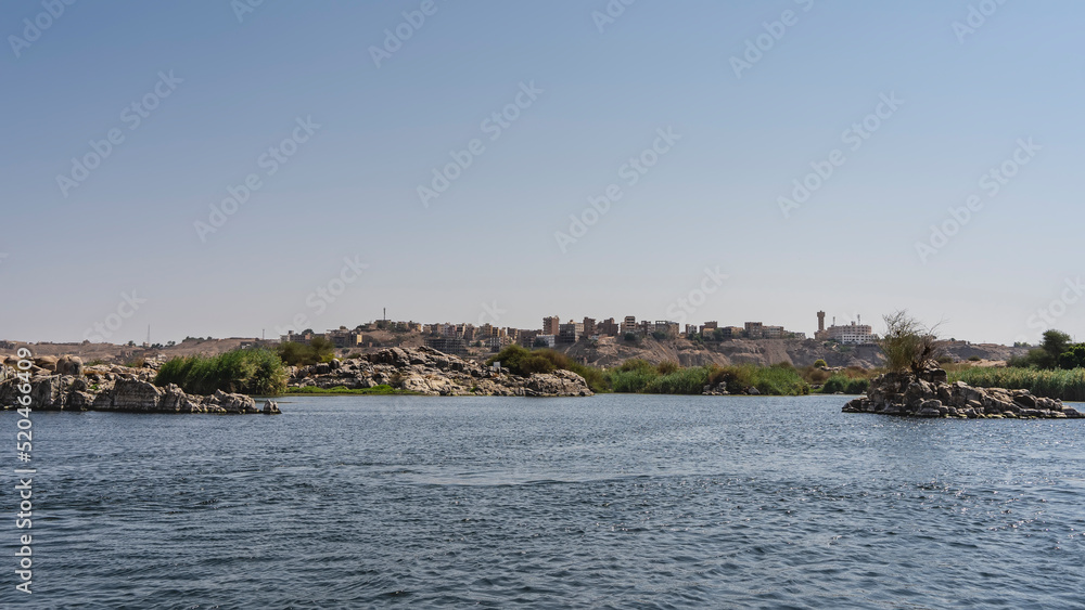 On the riverbank there are piles of boulders, green vegetation. Silhouettes of city buildings are visible against a clear sky. Ripples on the blue water. Copy space. Egypt. Nile