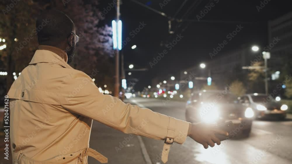 Slow motion shot of young adult African American man wearing stylish clothes and eyeglasses holding smartphone catching taxi late in evening Slow motion shot of young adult African American man wearing stylish clothes and eyeglasses holding smartphone catching taxi late in evening