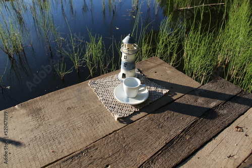 Lighthouse on wooden pier on riverside, empty white cup and soucer, forest flowers in sunny summer morning photo