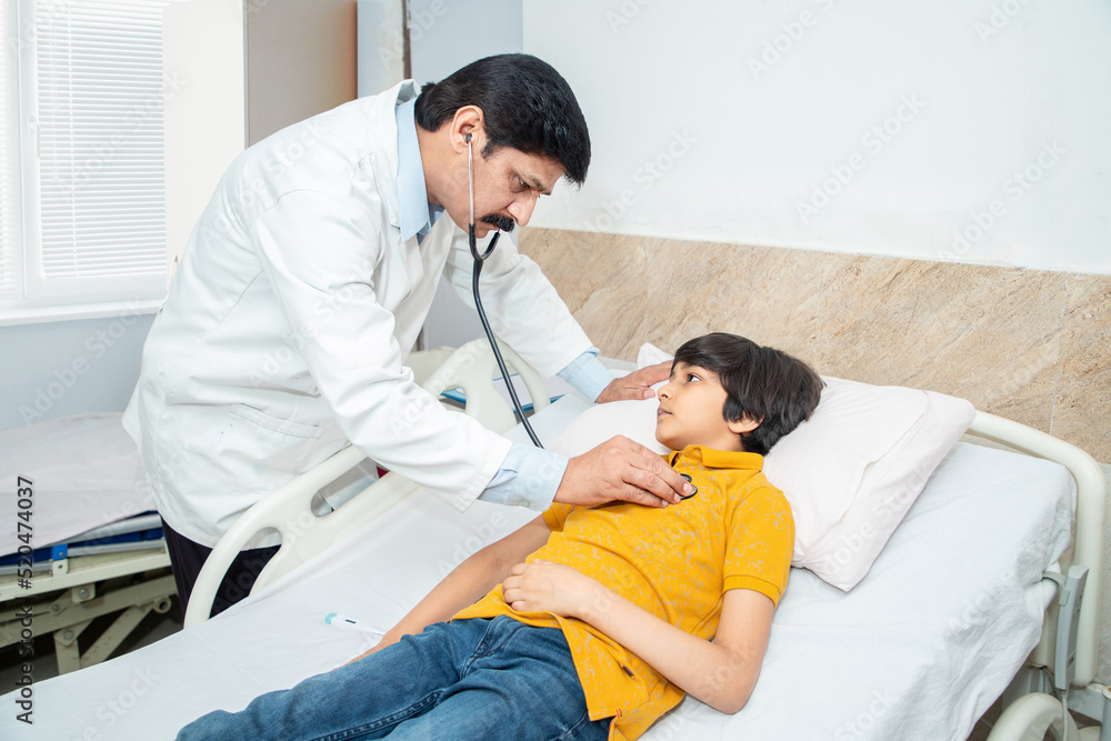 Obraz premium Indian doctor of pediatrician holding stethoscope checking heartbeat of sick boy kid at hospital. health checkup, children medical insurance care.