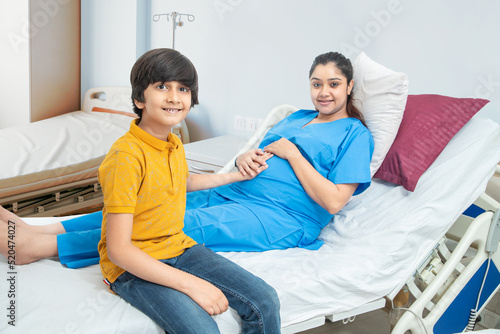 Happy indian son touching mother's pregnant belly at hospital or clinic bed. Happy asian boy feeling baby at mother tummy and awaiting the birth of his little brother.