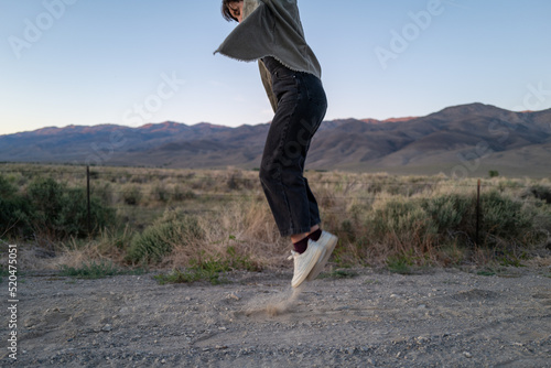 Short haired non-binary person jumps playfully on dusty road in open road landscape with mountains photo