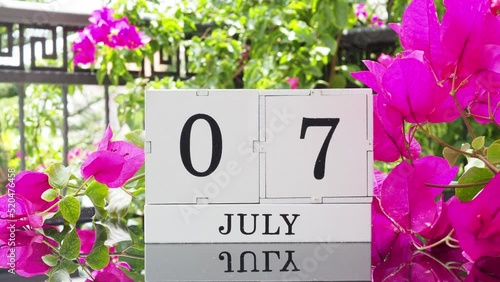 A wooden calendar with an important event for July 7 on a table with hibiscus flowers, a woman's hand sets the date on the calendar. Flip the calendar over to a cubic perpetual calendar. summer month photo