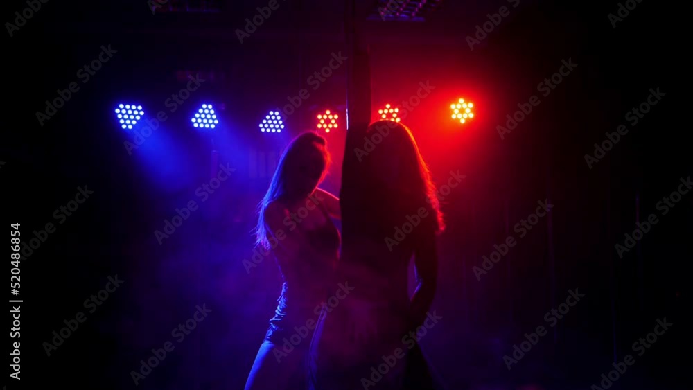 Two slender sexy women dance around a pole in the light of colorful lights with smoke in a strip club. Pole dance. Two slender sexy women dance around a pole in the light of colorful lights with smoke in a strip club. Pole dance.